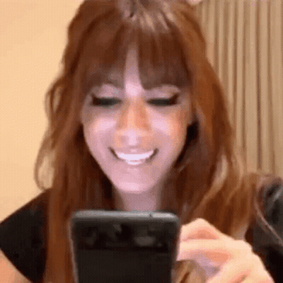 A Woman With Long Red Hair Is Smiling While Looking At Her Phone . GIF