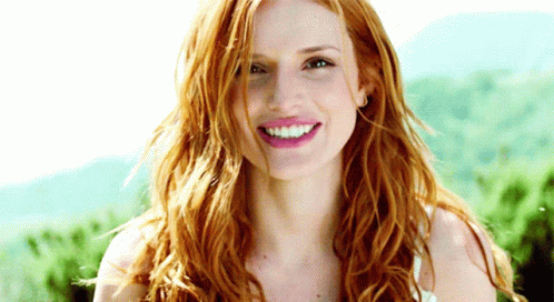 A Woman With Long Red Hair Is Smiling With Mountains In The Background GIF