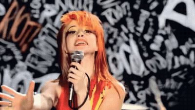 A Woman With Orange Hair Is Singing Into A Microphone In Front Of A Graffiti Wall . GIF