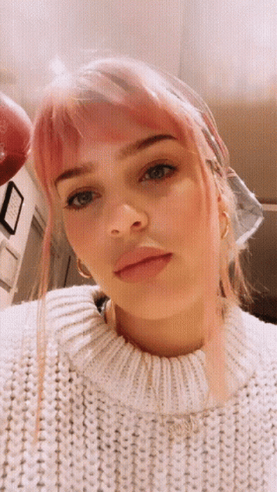 A Woman With Pink Hair And A White Sweater Looks At The Camera GIF