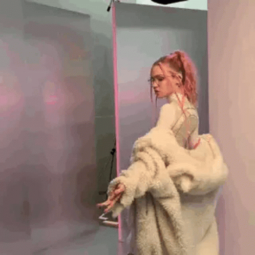 A Woman With Pink Hair And Glasses Is Wearing A Fur Coat And Standing In A Room . GIF