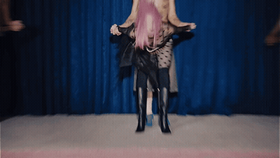 A Woman With Pink Hair Is Being Lifted On A Man 'S Shoulders In Front Of A Blue Curtain GIF