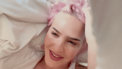 A Woman With Pink Hair Is Laying On A Bed And Smiling GIF