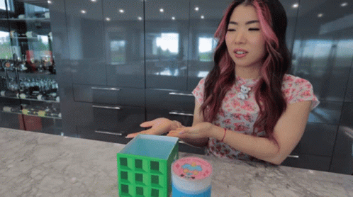 A Woman With Pink Hair Is Sitting At A Counter Holding A Container That Says ' Hello Kitty ' On It GIF