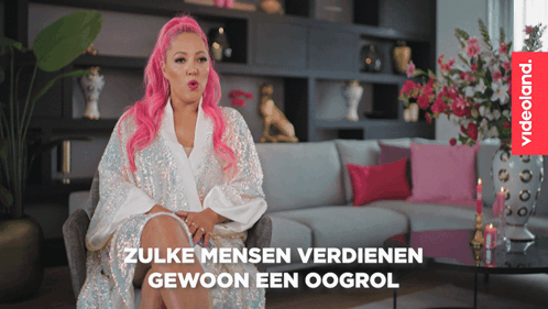 A Woman With Pink Hair Is Sitting In Front Of A Couch And A Videoland Logo GIF