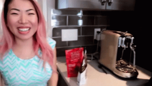 A Woman With Pink Hair Is Smiling In Front Of A Bag Of Coffee GIF