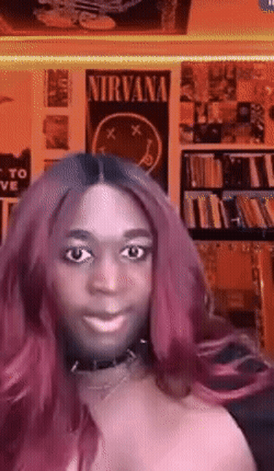 A Woman With Pink Hair Is Smiling In Front Of A Nirvana Poster On The Wall GIF