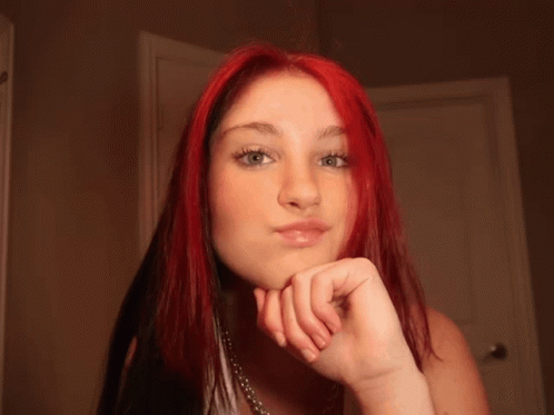 A Woman With Red Hair And Blue Eyes Looks At The Camera With Her Hand On Her Chin GIF