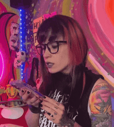 A Woman With Red Hair And Glasses Is Sitting At A Table Holding A Cell Phone . GIF