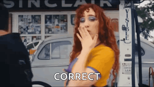A Woman With Red Hair Is Blowing A Kiss While Standing In Front Of A Gas Pump And A Car . GIF