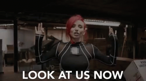 A Woman With Red Hair Is Making A Peace Sign And The Words Look At Us Now Are Behind Her GIF