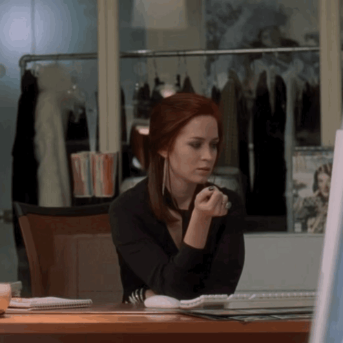 A Woman With Red Hair Is Sitting At A Desk Looking At A Computer Screen GIF