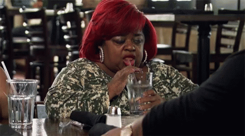 A Woman With Red Hair Is Sitting At A Table With A Glass Of Water And Eating Something . GIF