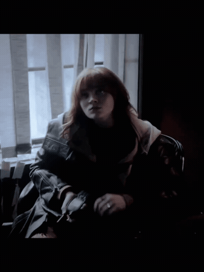 A Woman With Red Hair Is Sitting In Front Of A Window In A Dark Room GIF