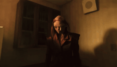 A Woman With Red Hair Is Standing In A Dark Room With A Fan On The Wall GIF
