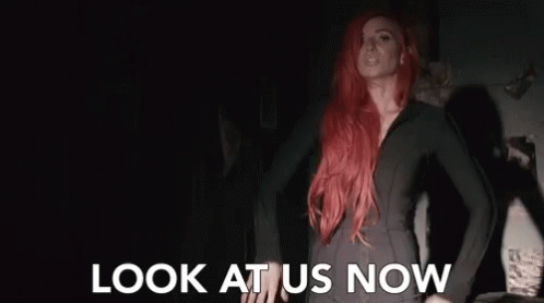A Woman With Red Hair Is Standing In A Dark Room With The Words Look At Us Now Above Her . GIF