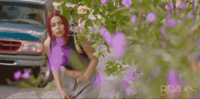 A Woman With Red Hair Is Standing In Front Of A Bush With Purple Flowers And The Word Bravis On The Bottom Right GIF