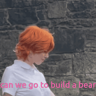 A Woman With Red Hair Is Standing In Front Of A Stone Wall With The Words " Can We Go To Build A Bear " Above Her GIF