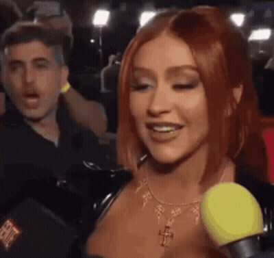 A Woman With Red Hair Is Talking Into A Microphone While A Man Looks On . GIF