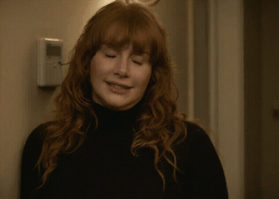 A Woman With Red Hair Is Waving Her Hand In Front Of A Door GIF
