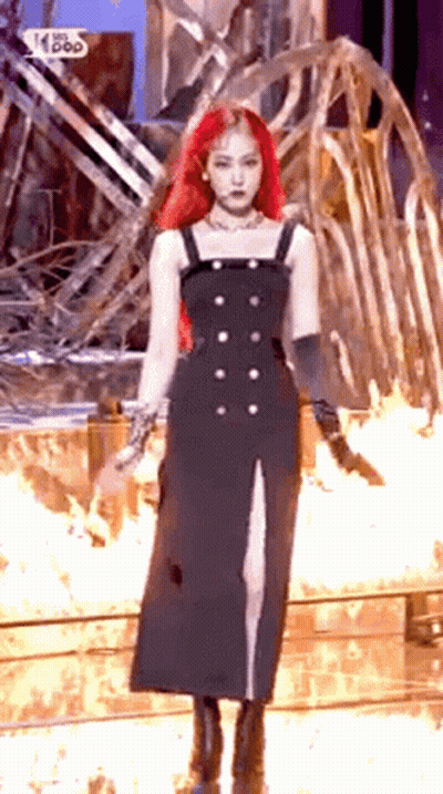 A Woman With Red Hair Is Wearing A Black Dress And Standing In Front Of A Fire . GIF