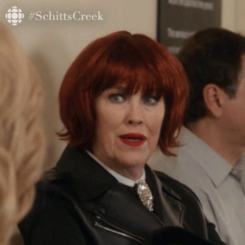 A Woman With Red Hair Is Wearing A Black Jacket And A #Schittscreek Logo Behind Her GIF
