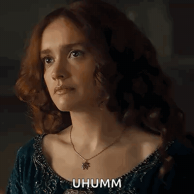 A Woman With Red Hair Is Wearing A Necklace And A Blue Dress And Looking At The Camera . GIF
