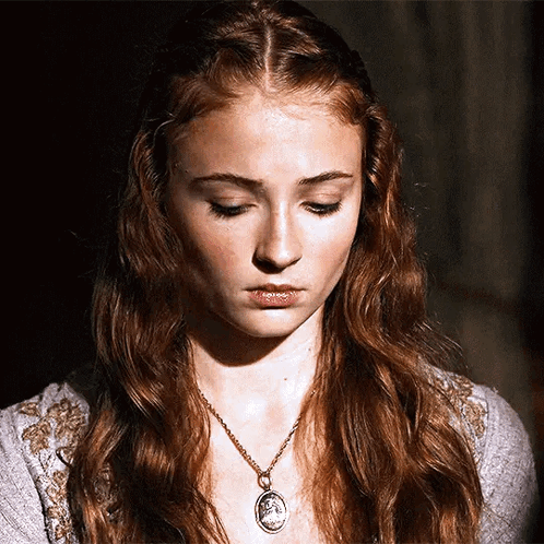 A Woman With Red Hair Is Wearing A Necklace With A Medallion On It GIF