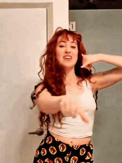 A Woman With Red Hair Is Wearing A White Tank Top And Black Pants GIF