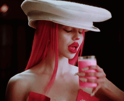 A Woman With Red Hair Wearing A White Hat And Holding A Glass Of Pink Liquid GIF