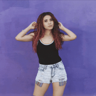 A Woman With Red Hair Wearing Shorts And A Black Tank Top Stands In Front Of A Purple Wall GIF