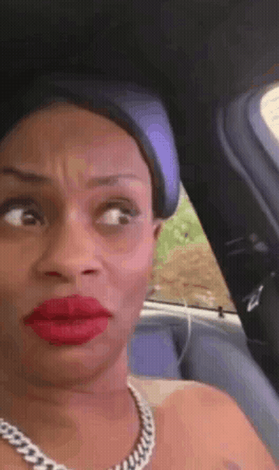 A Woman With Red Lipstick And A Necklace Is Sitting In A Car Making A Funny Face GIF