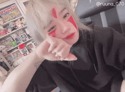 A Woman With Red Paint On Her Face Is Standing In Front Of A Shelf With A Monopoly Game GIF