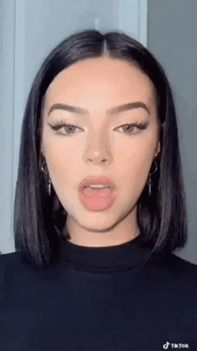 A Woman With Short Black Hair And Earrings Is Wearing A Black Sweater And Making A Funny Face . GIF