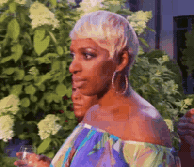 A Woman With Short Blonde Hair Is Holding A Glass Of Wine In Front Of A Bush . GIF