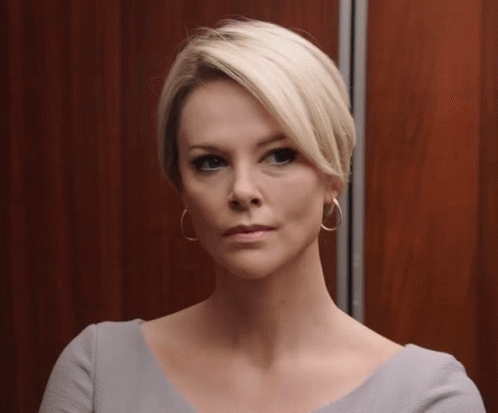 A Woman With Short Blonde Hair Wearing Hoop Earrings Looks At The Camera GIF