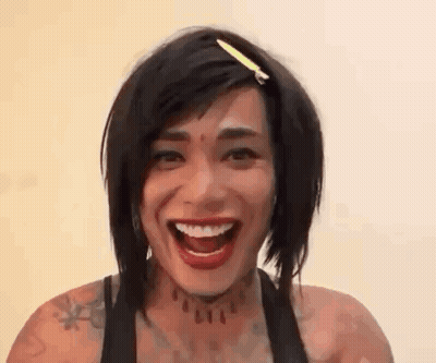 A Woman With Short Hair And A Tattoo On Her Neck Is Laughing With Her Mouth Open . GIF