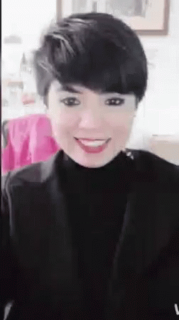 A Woman With Short Hair Is Smiling And Wearing A Black Turtleneck . GIF