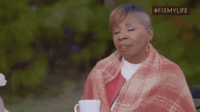 A Woman Wrapped In A Plaid Blanket Is Sitting At A Table With A Cup Of Coffee . GIF