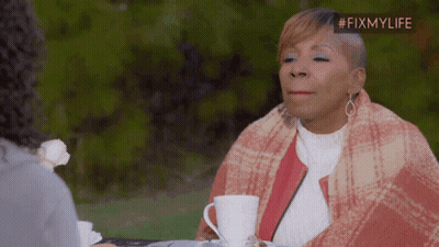 A Woman Wrapped In A Plaid Blanket Sits At A Table With A Cup Of Coffee In Front Of Her GIF