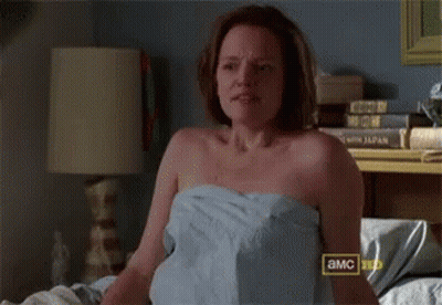 A Woman Wrapped In A Towel Is Sitting On A Bed With A Sign That Says A & Mc GIF
