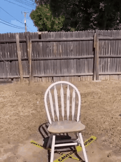 A Wooden Chair Sits In The Dirt In Front Of A Fence With A Yellow Caution Tape Around It GIF