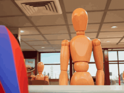 A Wooden Mannequin Sits At A Table In A Room GIF