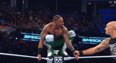 A Wrestler In A Green Outfit Holds Hands With Another Wrestler In Front Of A Smack Club Sign GIF