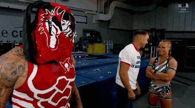 A Wrestler In A Red And White Outfit Is Talking To A Man And A Woman In A Room . GIF