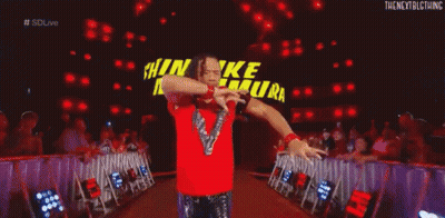 A Wrestler In A Red Shirt Is Kneeling Down In Front Of A Large Sign That Says Shinsuke Nakamura . GIF