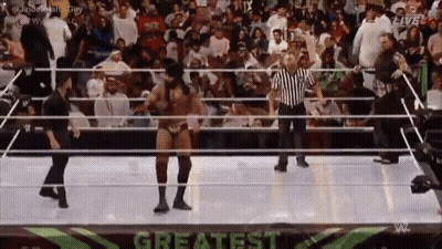 A Wrestler Is Dancing In A Wrestling Ring With A Referee In Front Of A Crowd . GIF