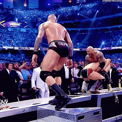 A Wrestler Is Jumping Over A Metal Box While Another Wrestler Is Kneeling Down . GIF