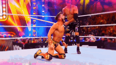 A Wrestler Is Kneeling Down In A Wrestling Ring While Another Wrestler Is Standing Behind Him . GIF