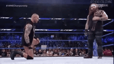 A Wrestler Is Kneeling Down In A Wrestling Ring While Another Wrestler Stands Behind Him . GIF